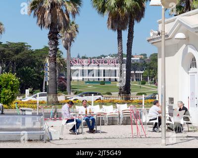 Estoril Casino hinter Palmen vom Bahnhof aus gesehen. Gemeinde Cascais, Portugal. Die Leute sitzen und trinken im Bahnhofscafé Stockfoto