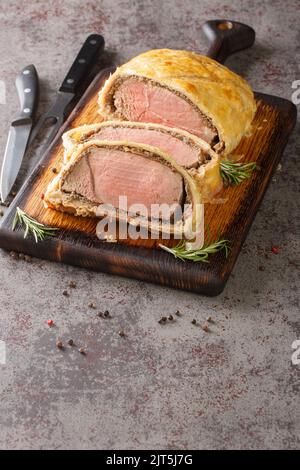 Geschnittenes Beef Wellington auf einem Holzbrett mit einem Rosmarinzweig auf grauem Hintergrund. Vertikal Stockfoto