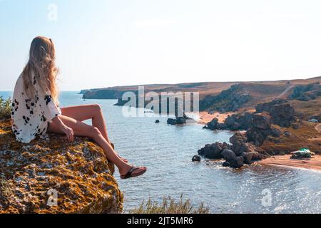 Eine Blondine mit langen Haaren sitzt auf einem Berg und blickt hinunter auf das Meer und die Felsen. Seascape, Reisen und Tourismus. Stockfoto