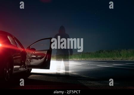 Eine geheimnisvolle, transparente, geisterhafte Figur, die auf einer gespenstischen leeren Straße neben einem Auto steht. In einer Sommernacht. Stockfoto