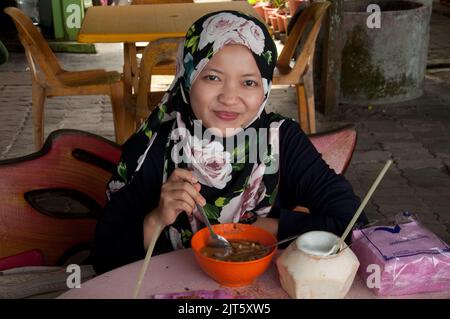 Junge muslimische Frau, die Laksa im Open-Air-Café Batu Ferringhi, Penang, Malaysia, Asien isst. Frisches Kokosnussgetränk; Laksa ist eine berühmte Suppe, die traditionell zubereitet wird Stockfoto