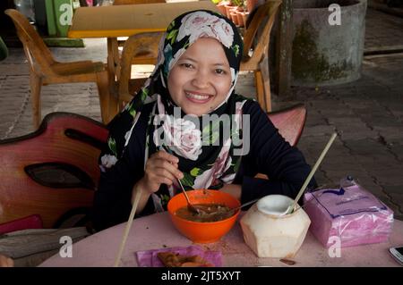 Junge muslimische Frau, die Laksa im Open-Air-Café Batu Ferringhi, Penang, Malaysia, Asien isst. Frisches Kokosnussgetränk; Laksa ist eine berühmte Suppe, die traditionell zubereitet wird Stockfoto