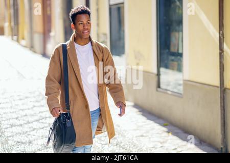 Ein junger schwarzer Mann, der die Straße entlang läuft, trägt eine Aktentasche und ein Smartphone. Stockfoto