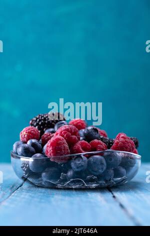 Eine Vielzahl von Beeren in Glaswaren auf türkisfarbenem Hintergrund. Himbeeren, Blaubeeren und Brombeeren in einer Schüssel. Stockfoto