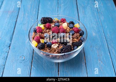 Eine Vielzahl von bunten Beeren und Müsli auf einem Teller. Glasplatte mit Granola und Beeren auf türkisfarbenem Hintergrund. Stockfoto