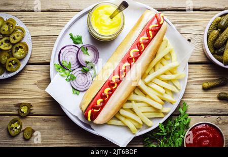 Hot Dog serviert mit Pommes frites, Gurkeneinchleinen, Jalapeno-Scheiben, frischer Zwiebel, frischer Petersilie, Honig-Senf und Ketchup. Draufsicht mit Nahaufnahme. Stockfoto