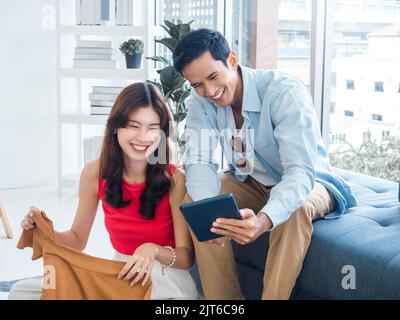 Glücklich schöner asiatischer Mann und junge schöne Frau lachen zusammen, während sie auf den Tablet-Bildschirm schauen. Frau hält ein Hemd und Mann hält Tablet auf Couc Stockfoto