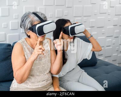 Asiatische alte ältere Frau, Mutter und glückliche junge Tochter, weiblich trägt VR-Brille, genießen sie 3D Spiel online zusammen im weißen Zimmer. Mutter und Tochter mit Stockfoto