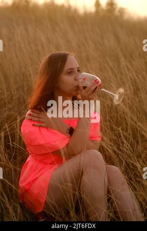 Junge Frau mit einem Glas Wein auf dem Feld. Seitenansicht eines jungen Weibchens in orangefarbenem Kleid, das Rotwein trinkt und wegschaut, während es auf Gras im Coun sitzt Stockfoto