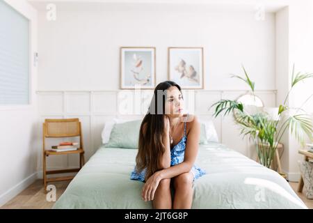 Traurige junge Erwachsene Frau, die auf dem Bett sitzt Stockfoto