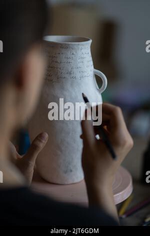 Weibliche Kunsthandwerkerin malt in der Werkstatt Text auf weiße Keramikvase mit Marker auf verschwommenem Hintergrund Stockfoto