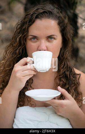 Wunderschöne weiße Frau mit lockigem, langem Haar und blauen Augen, die in eine bequeme Bettdecke oder einen gepolsterten Bettbezug eingewickelt ist, während sie in die Kamera schaut Stockfoto