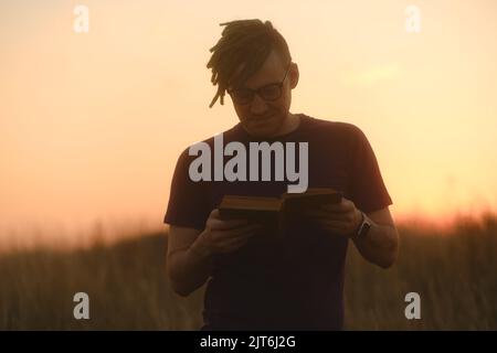 Lächelnder Hipster mit Buch. Niedriger Winkel des jungen Mannes mit Dreadlocks in legerer Kleidung und Brille, die mit einem Buch gegen den Sonnenuntergang steht. Stockfoto