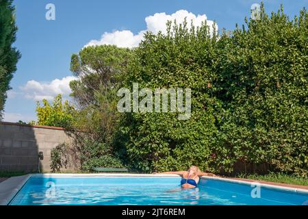 Eine blonde Frau entspannt sich in der Ecke eines Swimmingpools in einem Garten mit vielen Pflanzen Stockfoto