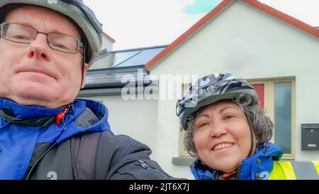 Nahaufnahme eines älteren Paares von Radfahrern, die Fahrradhelme tragen, bereit für eine Fahrradtour gegen ein verschwommenes Haus im Hintergrund, eine blaue Jacke und einen reflektierenden Ve Stockfoto
