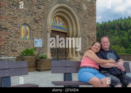 Lächelndes Touristenpaar mittleren Alters sitzt und umarmt auf Bank gegen Steinmauer und Eingang der Kirche Heiligen Cosmas und Damian, Bäume im Rücken Stockfoto