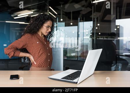 Schöne lateinamerikanische Geschäftsfrau, die in einem modernen Büro arbeitet, hat übermüdete Arbeitnehmerin starke Rückenschmerzen, die mit einem Laptop arbeiten Stockfoto