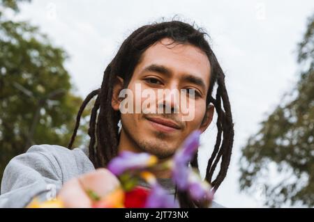 Porträt eines jungen venezolanischen Mannes mit Dreadlocks lächelnd und Blick auf die Kamera, im Freien mit der Hand Blumen, Konzept der Vielfalt und l Stockfoto