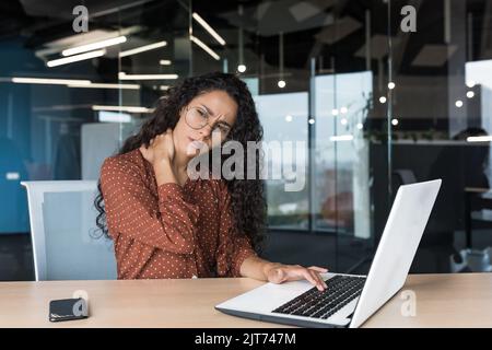 Übermüdete hispanische Frau, die im modernen Büro mit einem Laptop arbeitet, Geschäftsfrau im Bürogebäude, massiert Hals, Nackenschmerzen und Müdigkeit Stockfoto