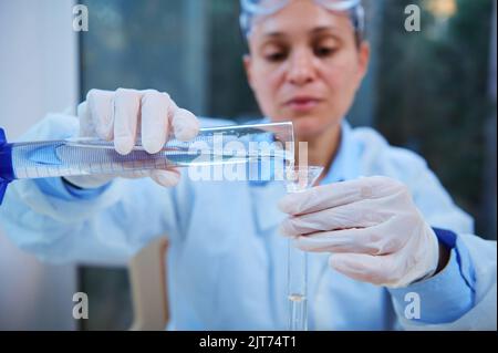 Details: Graduierter Zylinder in den Händen eines verschwommenen Wissenschaftlers, der Flüssigkeit durch den Labortrichter in das Reagenzglas gießt Stockfoto