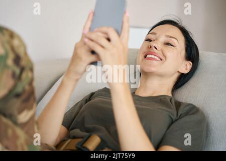 Militärfrau liegt auf dem Sofa, hält Handy in den Händen Stockfoto