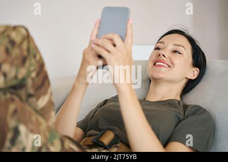 Soldatin liegt auf einer Couch und hält ein Mobiltelefon in den Händen Stockfoto