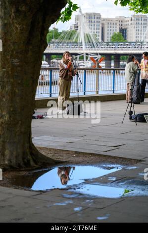 LONDON - 20. Mai 2022: Weibliche Straßenmusikerin spiegelte sich in der Pfütze am South Bank wider Stockfoto