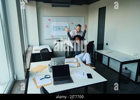 Mann im Büro schiebt freudige multirassische Frau in Sessel Stockfoto