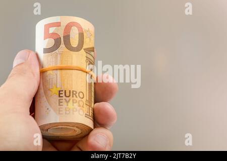 Die Hand hält eine Rolle von 50-Euro-Banknoten. Euro-Banknoten gerollt in der Hand eines weißen Mannes auf einem grauen Hintergrund. Das Konzept der finanziellen Unterstützung Stockfoto