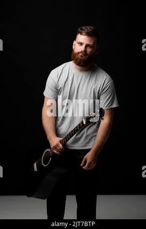 Der junge bärtige Gitarrist in grauem T-Shirt hält eine Gitarre in der Hand in einem dunklen Raum. Stockfoto