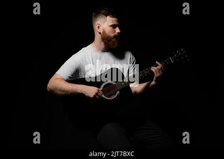 Der junge bärtige Gitarrist in grauem T-Shirt hält eine Gitarre und spielt in einem dunklen Raum ein Lied. Stockfoto