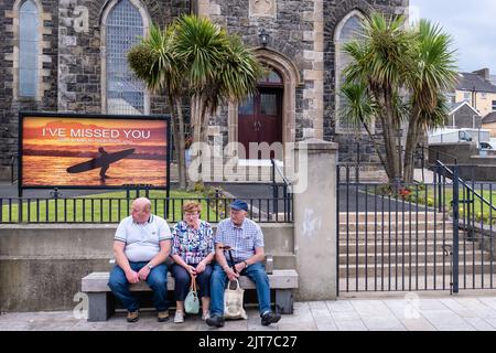 Drei Leute auf der Bank vor der Presbyterianischen Kirche. Das Hintergrundschild lautet: "Ich habe dich vermisst - Gott will von dir hören." Portrush, Großbritannien - 29. Juli 2022. Stockfoto