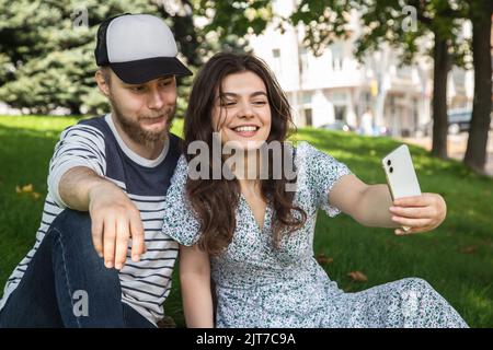 Ein Mann und ein Mädchen machen ein Selfie, das auf dem Gras im Park sitzt. Stockfoto