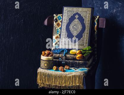 Islamisches Buch 'Quran' mit Tulpenblüten Rosenkranz, frischen Datteln und Gebetsteppich auf Holzstuhl platziert. Stockfoto