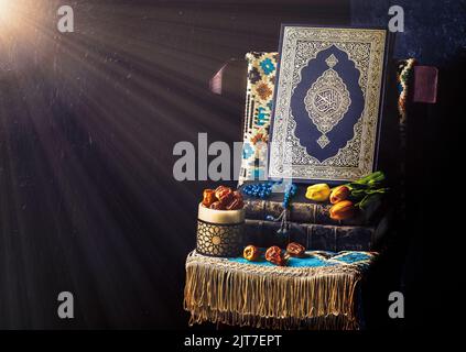 Das islamische Heilige Buch 'Quran' ist von Lichtstrahlen umgeben und mit Rosenkranz, frischen Datteln, Tulpenblumen und einem Gebetsteppich auf einem Holzstuhl platziert. Stockfoto