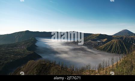 Mystischer und surrealer Panoramablick auf die Bromo Vulkankrater mit nebliger Oberfläche und grüner Vegetation, einschließlich des Dorfes Cemoro Lawang während des mornen Stockfoto