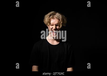 Wütender, trauriger, aggressiver junger Mann in einem schwarzen T-Shirt, der auf schwarzem Hintergrund schreit. Emotionaler Ausdruck. Stockfoto