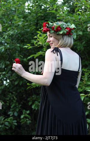 Eine stehende junge blonde Frau mit einem kurzen Haarschnitt in einem schwarzen langen Kleid und einem roten Kranz mit einer roten Blume vor grünen Bäumen, Seitenwand Stockfoto