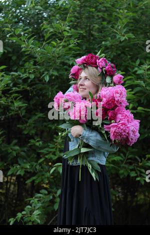 Eine stehende junge blonde Frau im Kranz, die im Sommer einen Strauß rosa Blumen vor grünen Bäumen hält und in den Himmel blickt, mit einem Seitengesicht Stockfoto