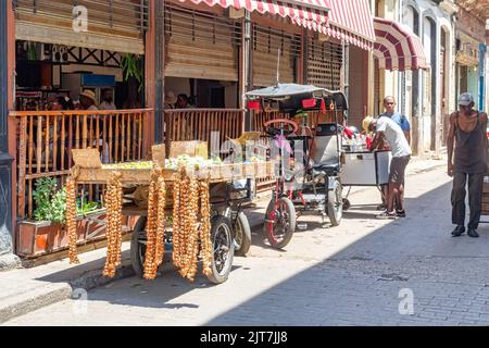 In einer engen Stadtstraße in der Altstadt von Havanna steht ein Lebensmittelkarren. Andere kleine Unternehmen stehen im Hintergrund. Ein schlanker Mann läuft in der Gegend. Stockfoto