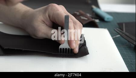 Nahaufnahme Herrenhände machen ein Lederprodukt. Löcher in das Leder mit einem Locheisen mit einem Hammer bohren. Werkzeuge für die Lederhandwerk. Stockfoto