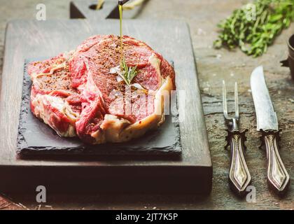 Gießen von nativem Olivenöl auf frisches rohes Rindersteak mit Gewürzen, Butter und Rosmarin. Nahaufnahme des Kopierbereichs. Stockfoto