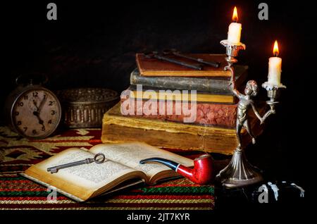 Klassisches Stillleben mit alten Büchern, die mit alter Uhr, antikem Silberkasten, Pfeife, alten Schlüsseln und beleuchteten Kerzen in kupfernen antiken Kerzenleuchtern platziert sind. Stockfoto