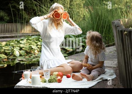 Porträt einer fröhlichen Familie von Mutter und kleiner Tochter, die auf Karli sitzt. Junge Frau, die Grapefruit in der Nähe der Augen hält. Stockfoto