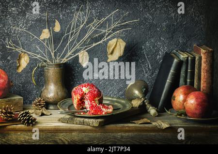 Klassisches Stillleben mit frischen Granatäpfeln, die mit Vintage-Büchern, antiken Kerzenständern und trockenen Herbstblättern in altem Kupferstich platziert sind. Stockfoto