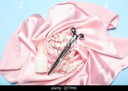 Gesichtsmassagewerkzeug mit Kosmetikflasche, Blumen und Stoff auf blauem Hintergrund Stockfoto