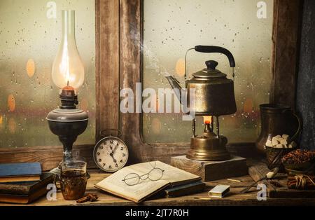 Klassisches Stillleben mit heißer Teeschale, die mit beleuchteter Vintage-Lampe, alten Büchern, einer Tasse Tee auf einem rustikalen Holztisch platziert ist. Stockfoto