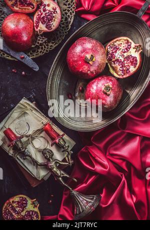 Klassisches Stillleben mit frischen Granatäpfeln, platziert mit Vintage-rotem Kerzenständer und Seidenstoff auf dunklem Hintergrund. Stockfoto