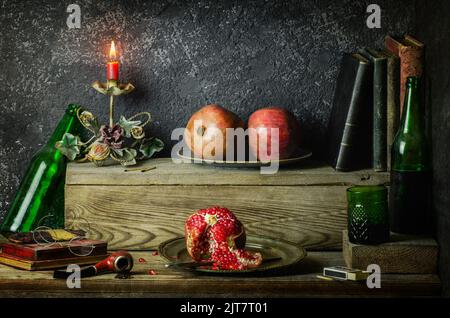 Klassisches Stillleben mit frischen Granatäpfeln, die mit alten Büchern, antiken Kerzenständern, Getränken, alter Pfeife und Gläsern auf rustikalem Holzhintergrund platziert sind. Stockfoto