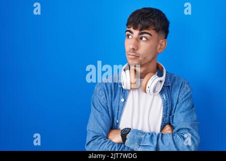 Junger hispanischer Mann, der auf blauem Hintergrund steht und mit überkreuzten Armen zur Seite blickt, überzeugt und zuversichtlich Stockfoto